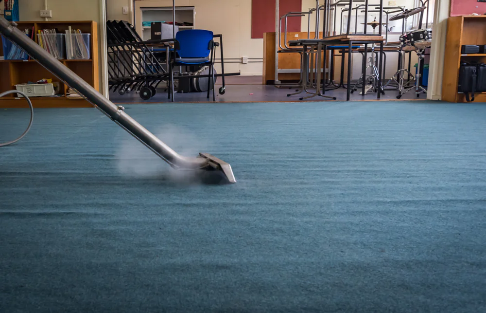 A carpet cleaning machine steams a blue carpet in an empty classroom, with stacked chairs, shelves, and a blue chair visible in the background.