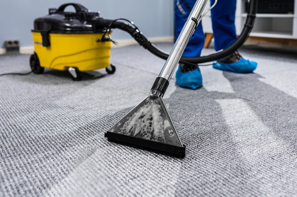 A person wearing blue shoe covers uses a carpet cleaning machine with a yellow vacuum to clean a gray carpet in a room.