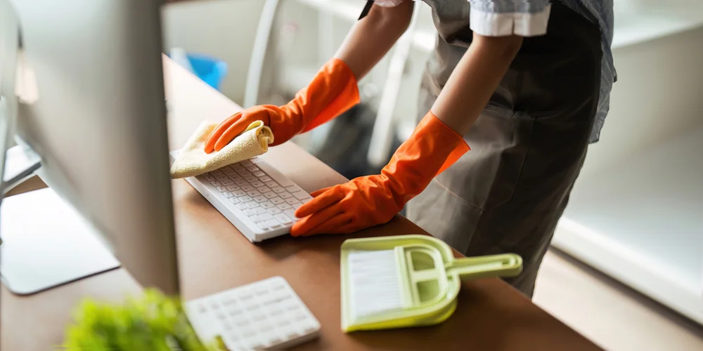 Person wearing orange gloves cleaning a computer keyboard with a yellow cloth; a dustpan and brush are on the desk nearby. The workspace is neat and well-lit.