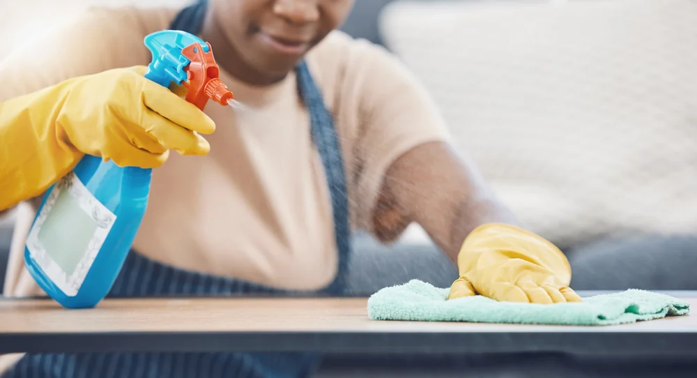 A person wearing yellow rubber gloves sprays cleaning solution onto a wooden surface and wipes it with a green cloth.