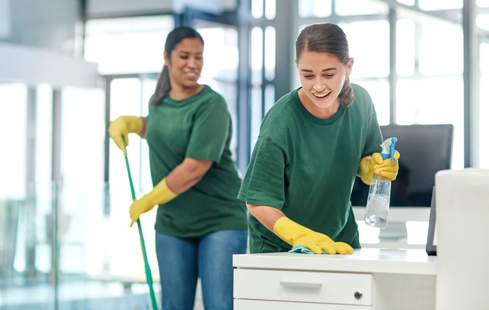 Two women in green shirts and yellow gloves clean an office. One woman wipes a desk with a cloth and spray bottle, while the other mops the floor. Both are smiling and there is natural light coming through the windows.