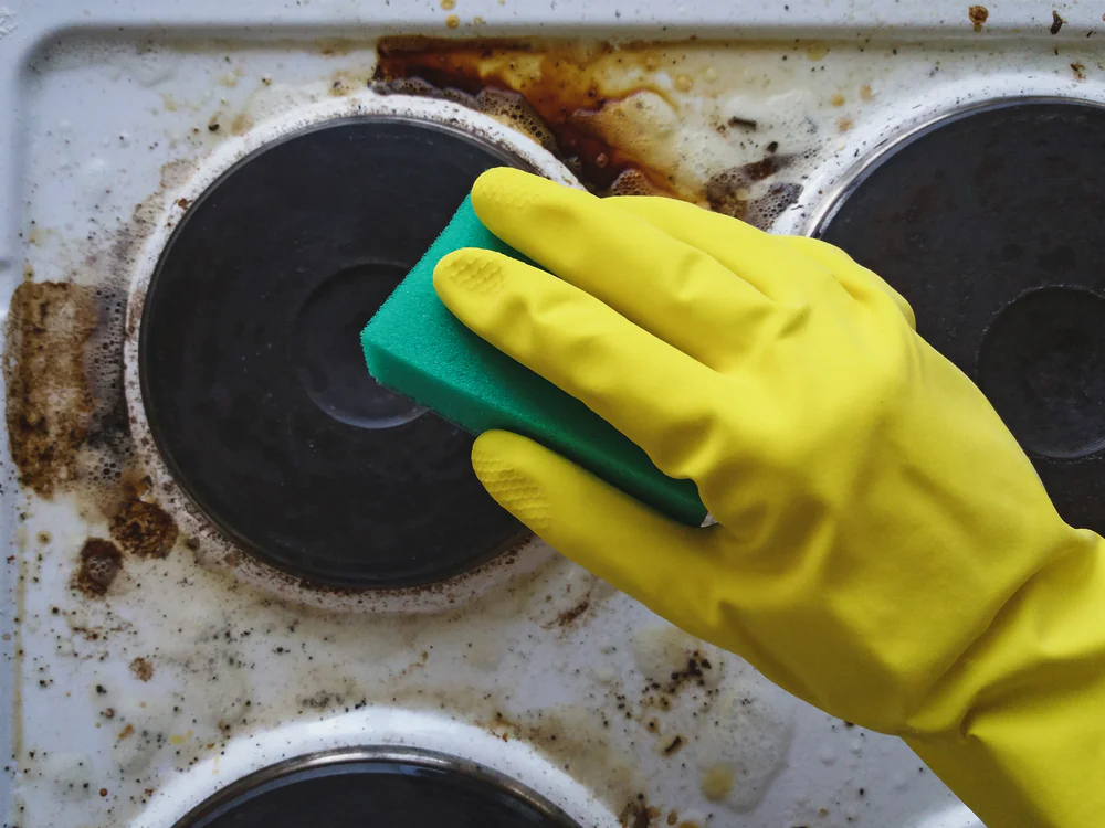 A hand wearing a yellow rubber glove holds a green sponge, scrubbing a dirty stovetop with burnt-on stains and grime.