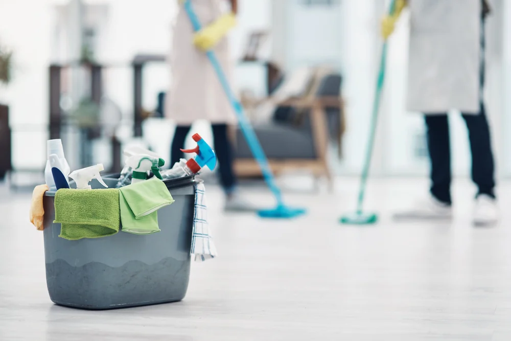 A bucket filled with cleaning supplies sits on the floor, while two people in the background, wearing gloves and aprons, mop a bright, modern room.