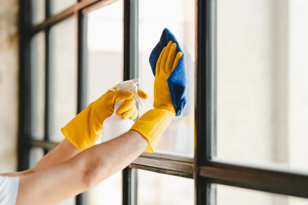 A person wearing yellow rubber gloves cleans a window using a blue cloth and a spray bottle, wiping the glass from the inside.