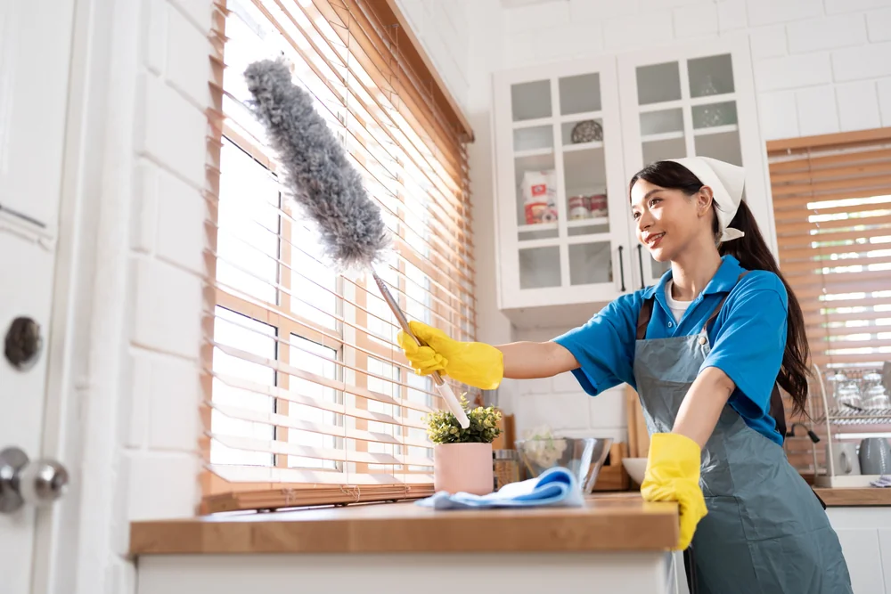 A woman wearing yellow gloves and a blue shirt dusts window blinds in a bright kitchen. She is smiling and using a fluffy duster, with cleaning supplies and a small plant on the counter nearby.