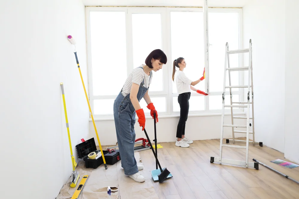 Two women in work clothes and rubber gloves clean and renovate a bright room; one sweeps the floor while the other paints near a large window. A ladder and tools are visible nearby.