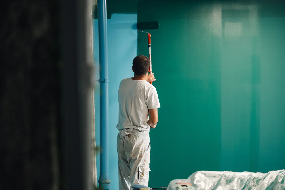 A person dressed in white painting a wall teal with a roller brush; some furniture is covered with plastic sheets nearby.