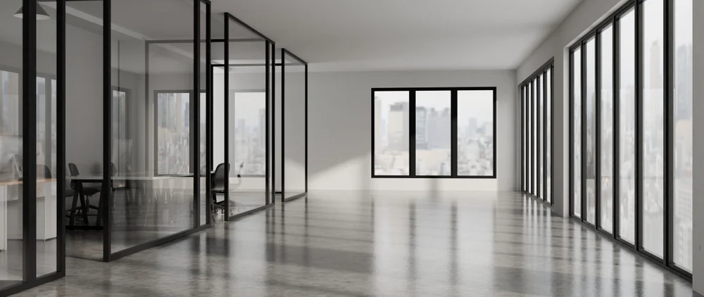 A modern, empty office space with large floor-to-ceiling windows, glass-walled meeting rooms, and a polished concrete floor. City skyline visible through the windows.