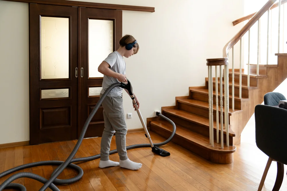 A person wearing headphones vacuums a wooden floor near a staircase in a bright, modern home. They are dressed casually and appear focused on cleaning.