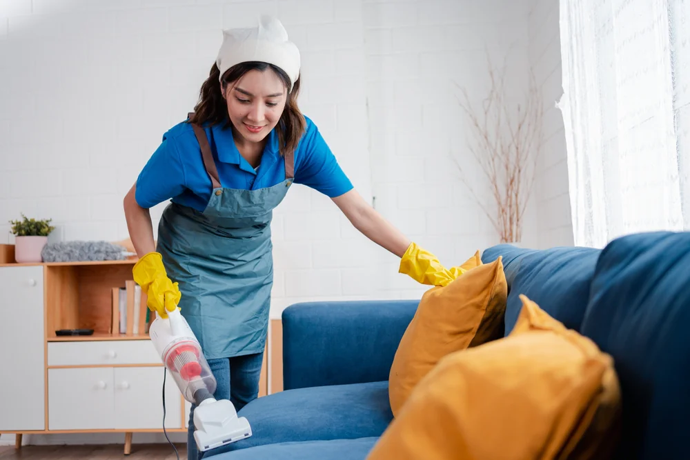 A woman wearing a headscarf, apron, and yellow gloves is smiling while vacuuming a blue sofa with yellow cushions in a brightly lit living room.